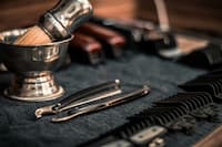 Close-up of barber tools including a shaving brush, straight razor, and hair clipper attachments on a dark cloth